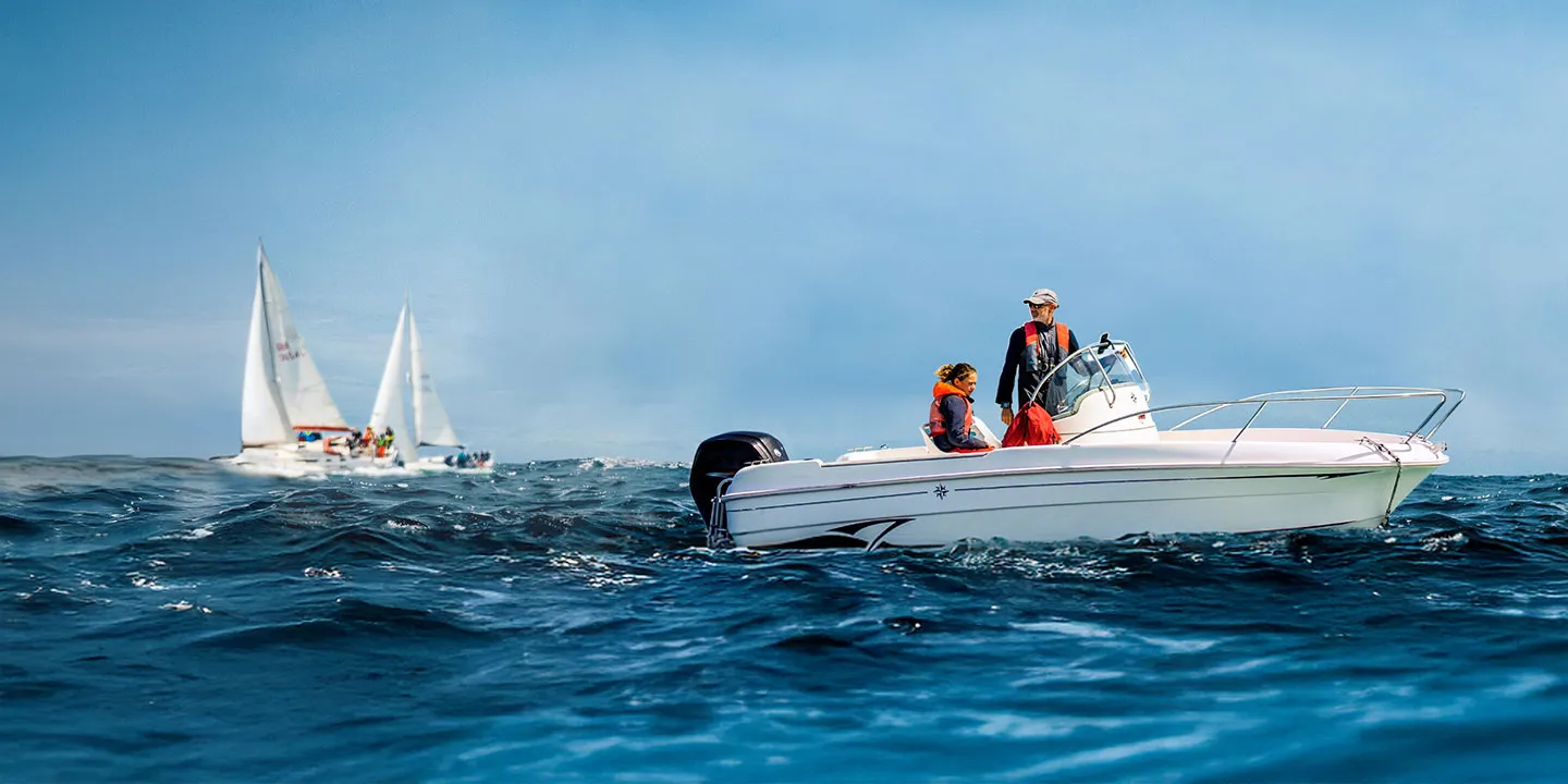 A small white pleasure vessel on open sea. In the background, sailing vessels can be seen on the water. HM Coastguard's recreational boating safety campaign image