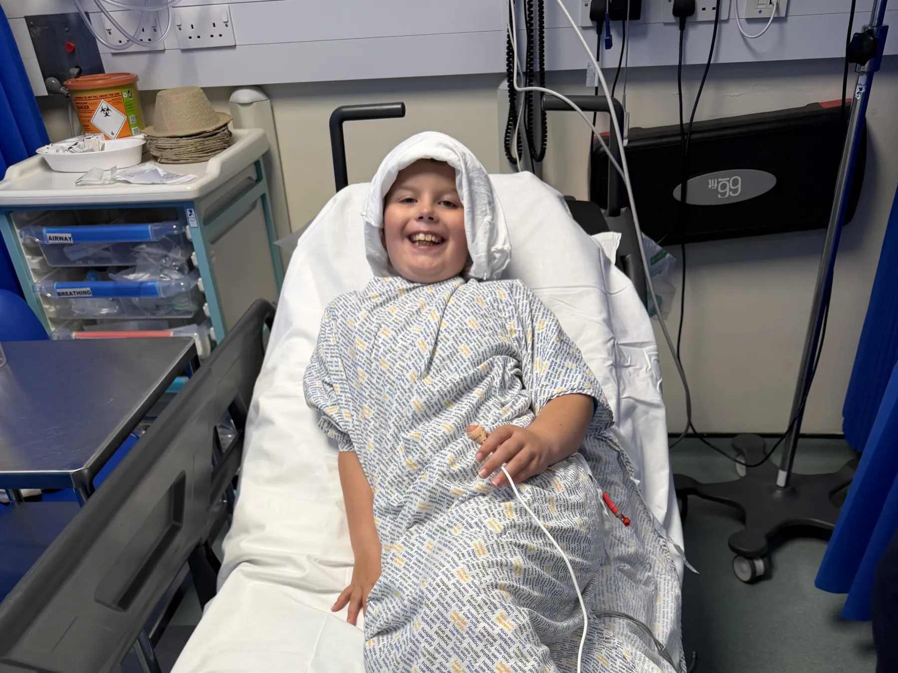 Clayton in a patient's gown smiling as he lays on a hospital bed