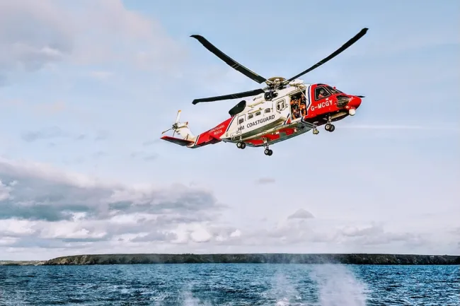 A white and red HM Coastguard helicopter hovers low in the sky above the sea with the coastline in the background.