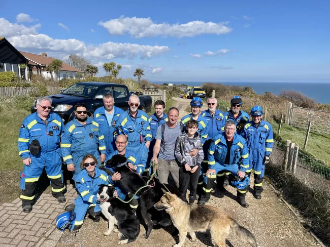 HM Coastguard volunteer coastguard rescue officers stood with the father, son and dogs rescued. In the distance, a coastguard rescue vehicle and the ocean are visible. 