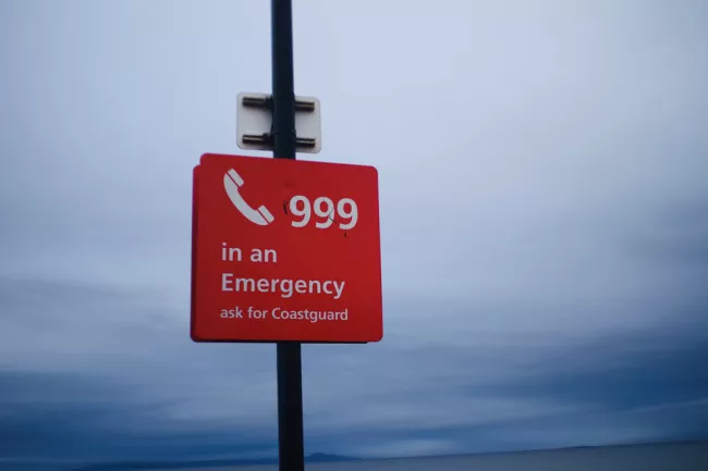 Red sign with 'call 999 in an emergency, ask for the Coastguard' with grey sky backdrop