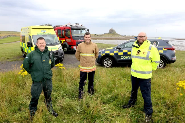 North East Ambulance Service, Fire and Rescue Service and HM Coastguard representatives stand in front of their vehicles with Holy Island in the background