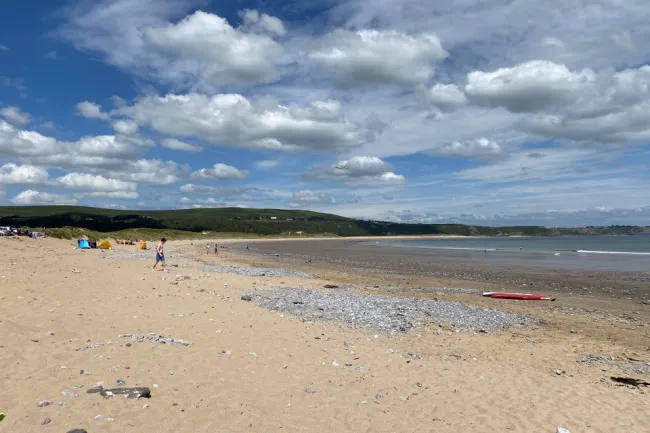 A view of Oxwich Bay with a blue and cloudy sky giving way to a sandy beach with slight pebbling at low tide. 