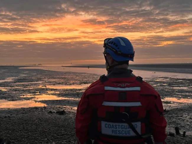 A member of the Southend Coastguard Rescue Team stands on the shoreline amid a golden sunset and low tide. 
