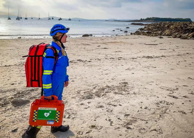 A Coastguard Rescue Officer, in blue uniform and helmet, walks along sands on the UK coastline.