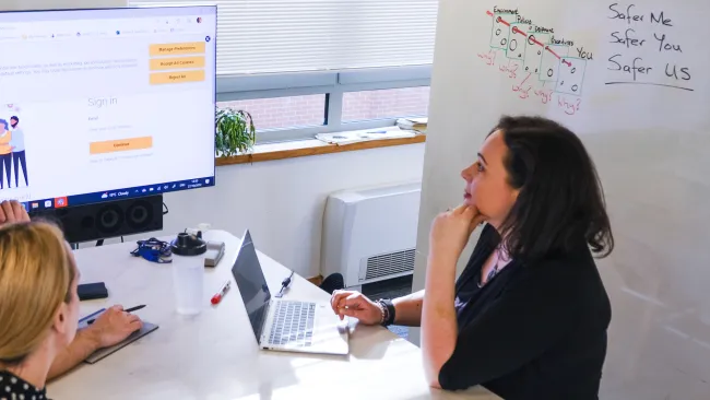 Two office workers on either side of a desk contemplate a screen