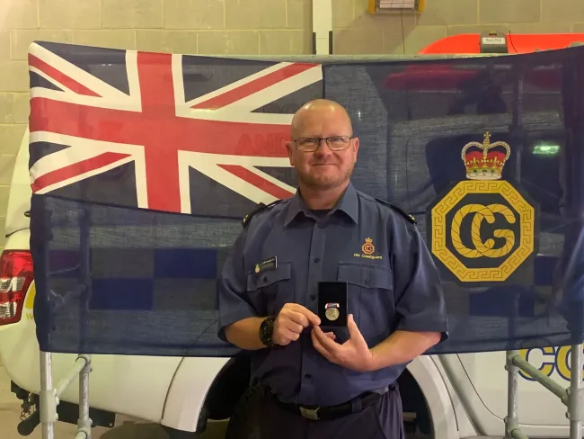 Coastguard Rescue Officer Kevin Moran in uniform stands in front of an HM Coastguard flag