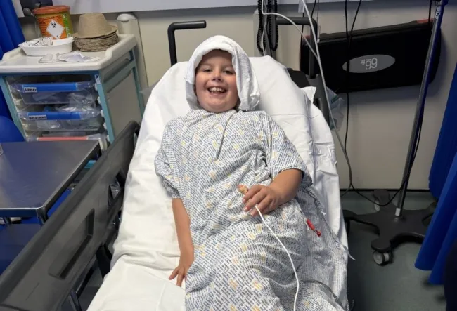 Clayton in a patient's gown smiling as he lays on a hospital bed