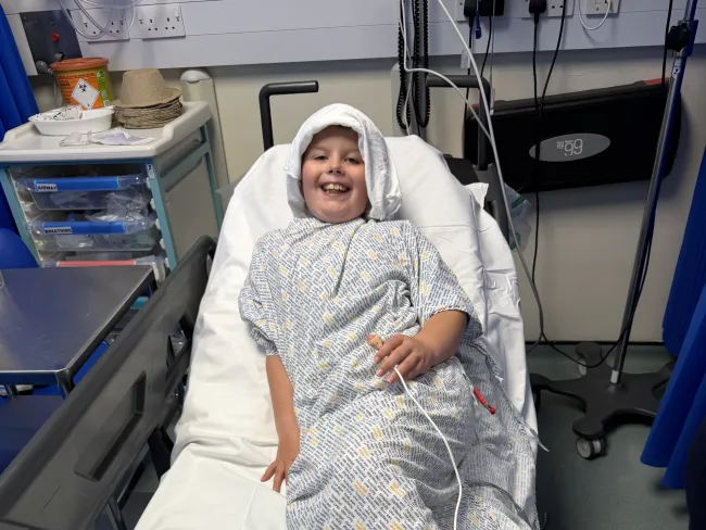 Clayton in a patient's gown smiling as he lays on a hospital bed