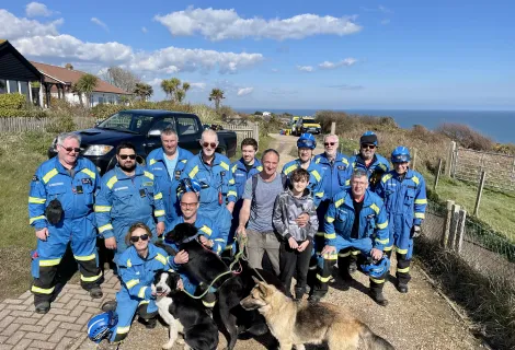 HM Coastguard volunteer coastguard rescue officers stood with the father, son and dogs rescued. In the distance, a coastguard rescue vehicle and the ocean are visible. 