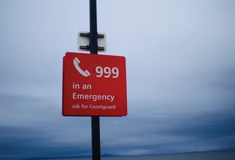 Red sign with 'call 999 in an emergency, ask for the Coastguard' with grey sky backdrop