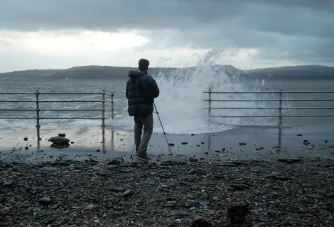 Man with tripod on harbour wall with water spray in stormy conditions