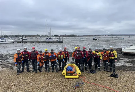 Members of HM Coastguard and Hampshire and Isle of Wight Fire and Rescue Service smiling for a group photo amidst sand and mud in Eastney, Portsmouth, equipped with mud rescue kit
