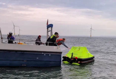 A luminous life raft is collected from the sea by boat crew with offshore wind turbines in the background