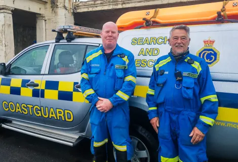 HM Coastguard Fleetwood Coastguard Rescue Team members Steve and Alan stand in blue emergency boiler suits beside the team's Coastguard Rescue Vehicle. 