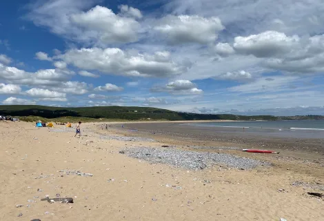 A view of Oxwich Bay with a blue and cloudy sky giving way to a sandy beach with slight pebbling at low tide. 