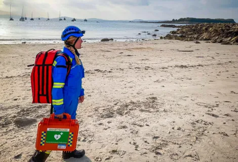 A Coastguard Rescue Officer, in blue uniform and helmet, walks along sands on the UK coastline.