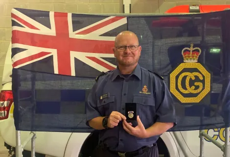 Coastguard Rescue Officer Kevin Moran in uniform stands in front of an HM Coastguard flag