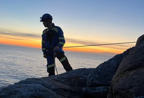 A coastguard roped to a cliff looks out to sea silhouetted by a sunset