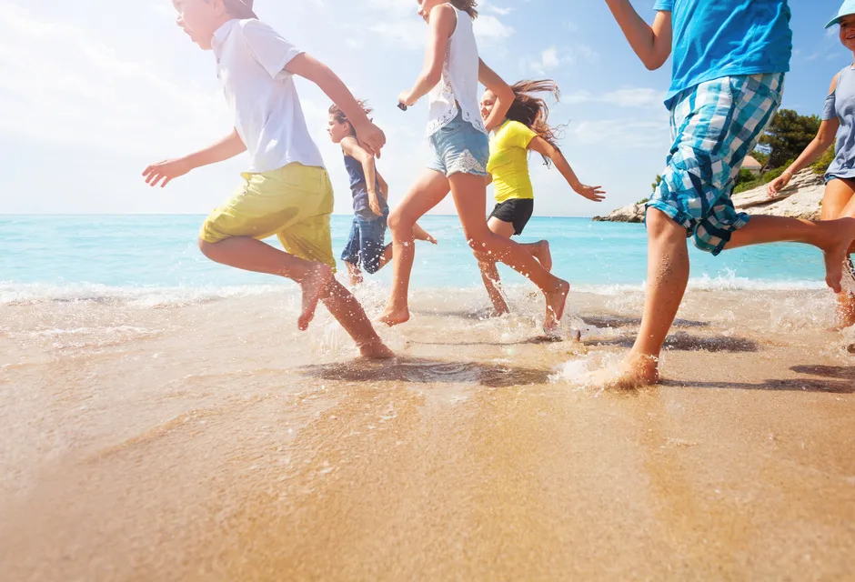 Picture of six children running on sand, focus on legs, near water's edge on sunny day