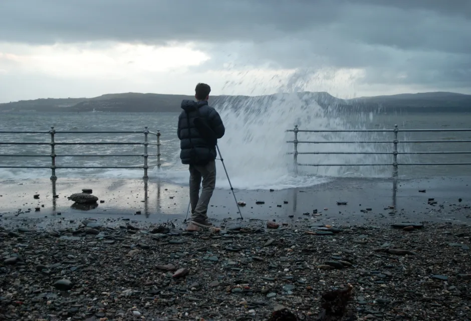 Man with tripod on harbour wall with water spray in stormy conditions