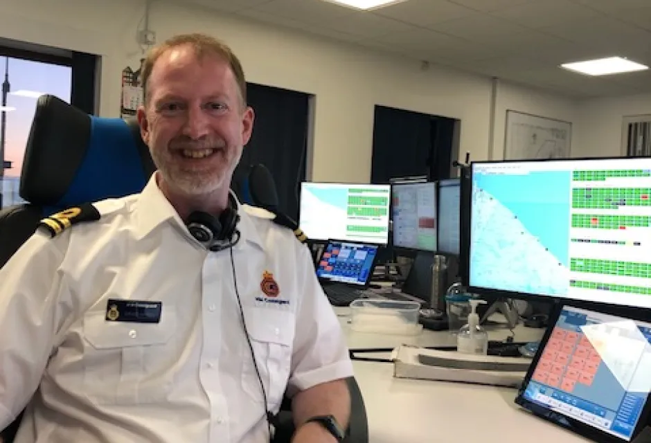 Smiling man in Coastguard uniform in front of bank of computer screens