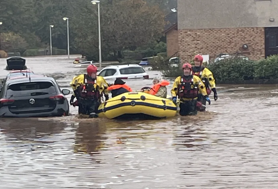 Four men in high vis and with flood rescue equipment wade through waist high water in residential street as they pass a part submerged car