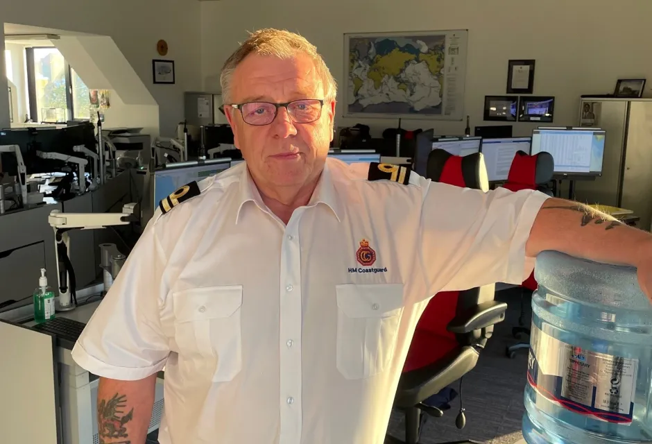 Unsmiling man in Coastguard uniform stood in front of camera in operations room.