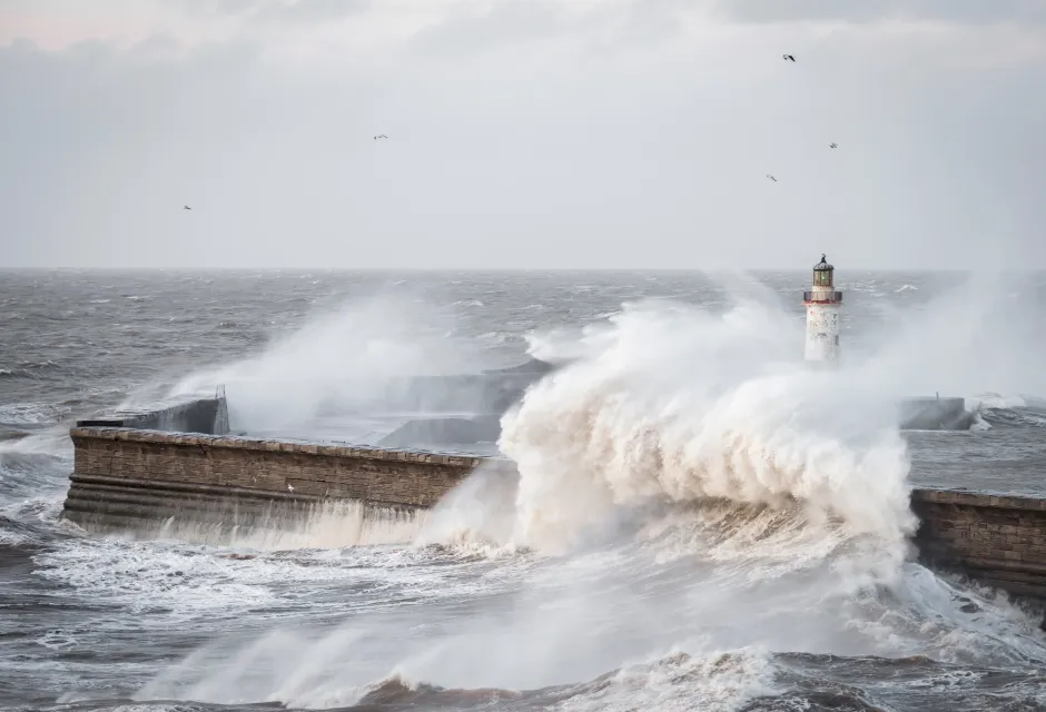 Storm waves crash over harbour wall and lighthouse in stormy seas