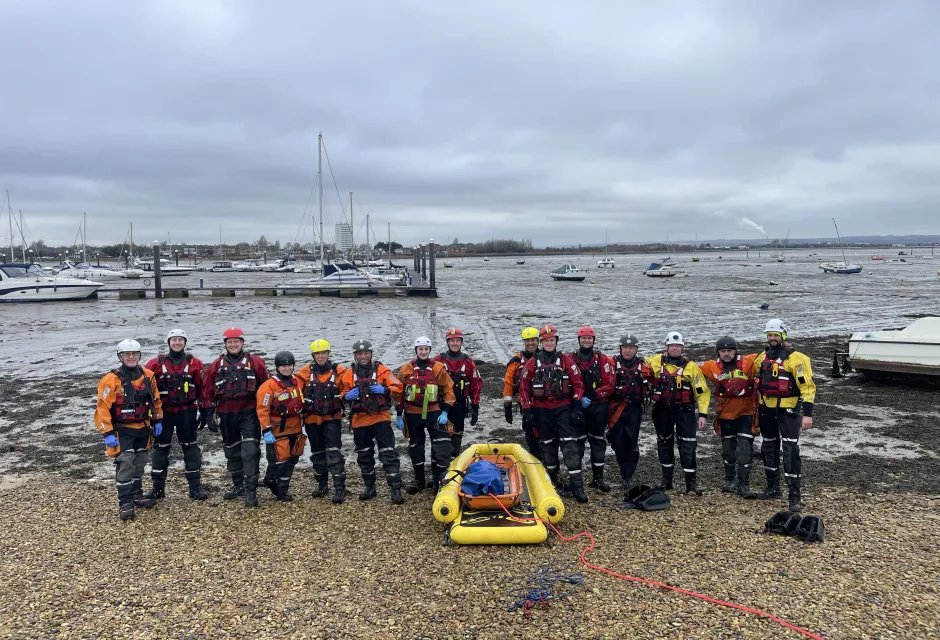 Members of HM Coastguard and Hampshire and Isle of Wight Fire and Rescue Service smiling for a group photo amidst sand and mud in Eastney, Portsmouth, equipped with mud rescue kit