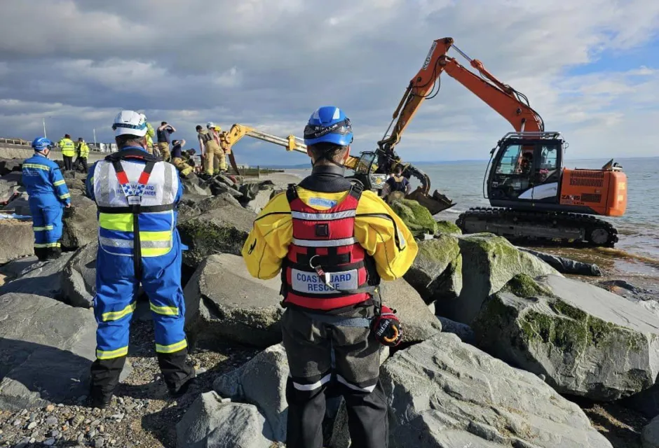 Coastguard officers observe as an excavator operates by rock armour at Tywyn beach