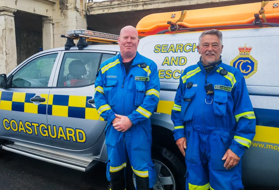 HM Coastguard Fleetwood Coastguard Rescue Team members Steve and Alan stand in blue emergency boiler suits beside the team's Coastguard Rescue Vehicle. 