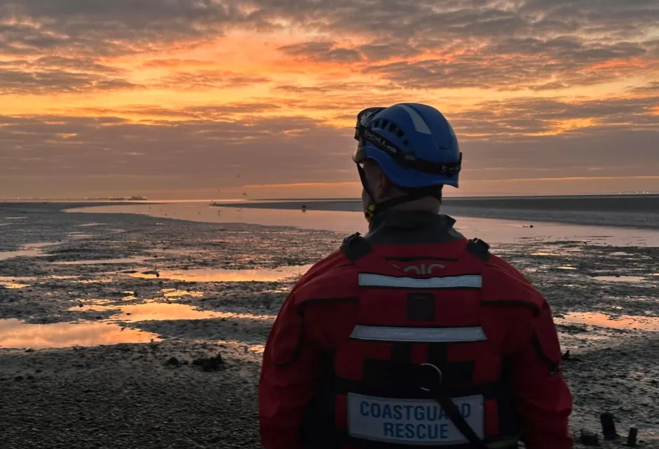 A member of the Southend Coastguard Rescue Team stands on the shoreline amid a golden sunset and low tide. 