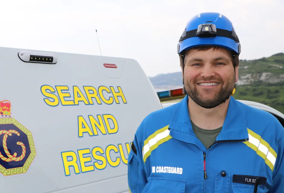 Tom Nunnen-Wills, wearing blue HM Coastguard overalls, standing in front of an HM Coastguard vehicle