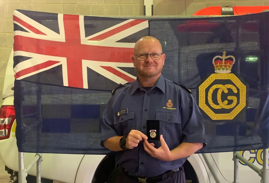 Coastguard Rescue Officer Kevin Moran in uniform stands in front of an HM Coastguard flag