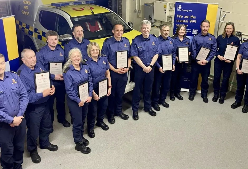 Smiling Coastguards in blue uniforms hold their awards with Assistant Chief Coastguard John Craig standing in the middle